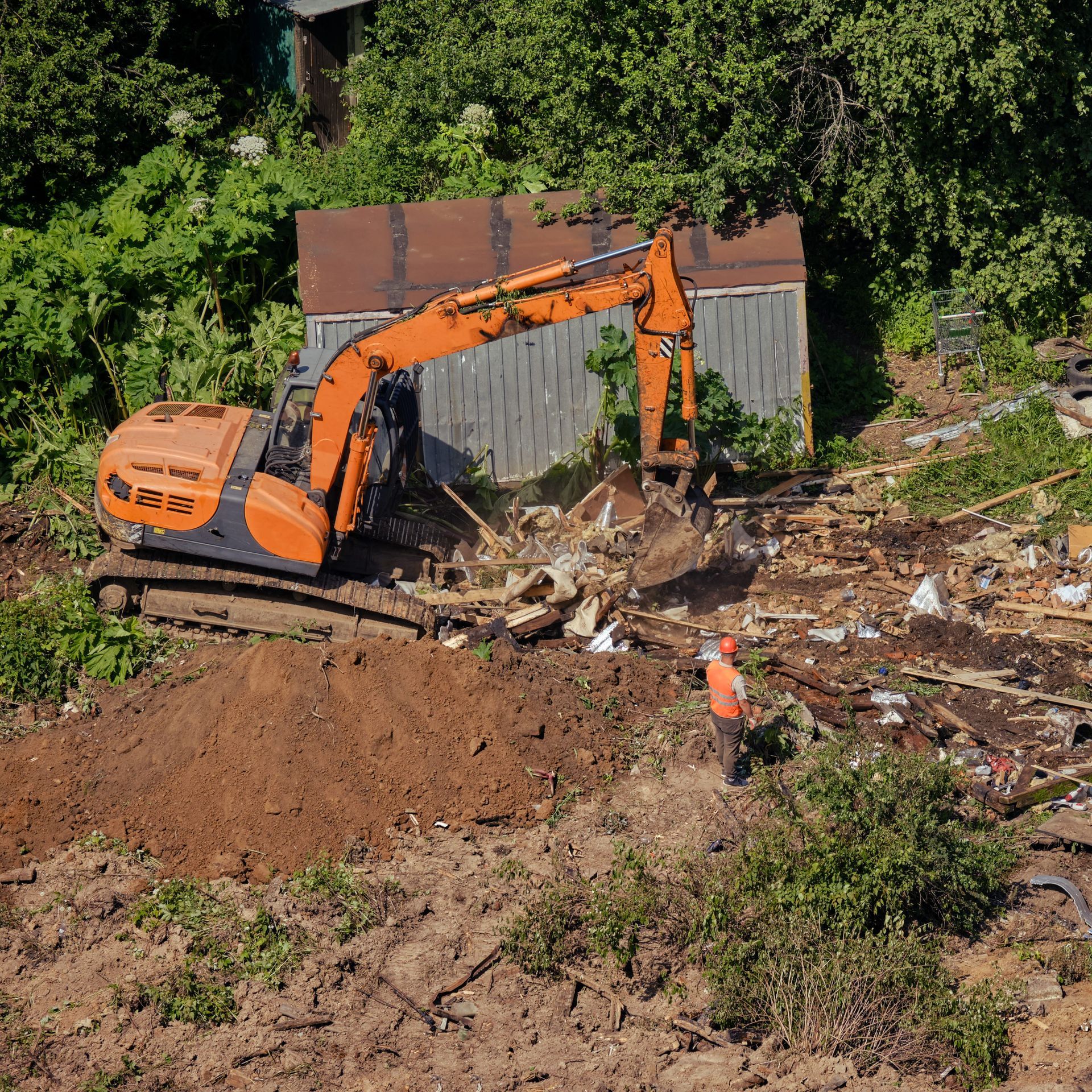 An orange excavator demolishes a building with a worker nearby in a forested area.
