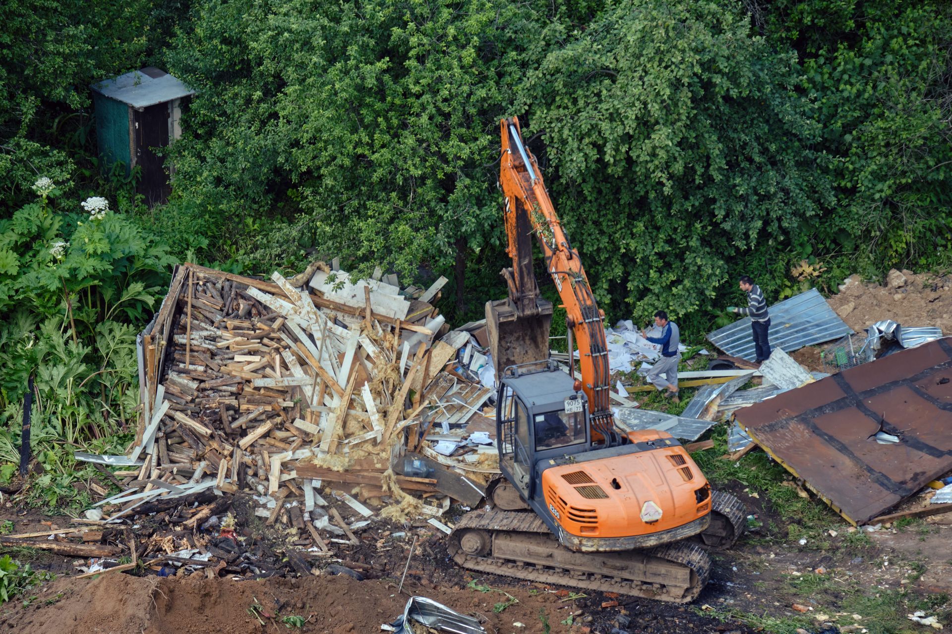 An excavator demolishes a wooden structure, with debris scattered. Green trees surround the area, with a small outhouse nearby.