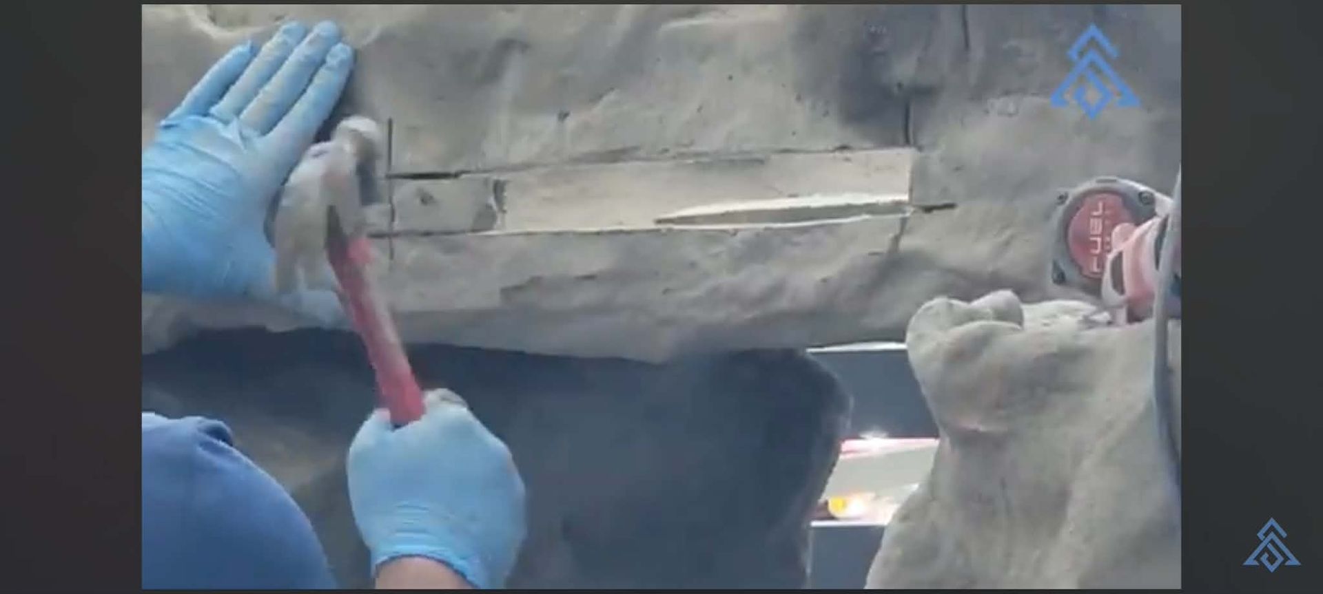 Gloved hands using a hammer and chisel on a stone wall, with dust and a small bucket nearby