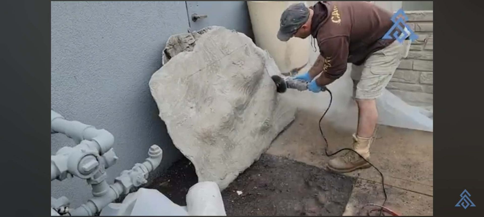 Worker using a power tool to cut a large stone slab outdoors beside a wall and pipes