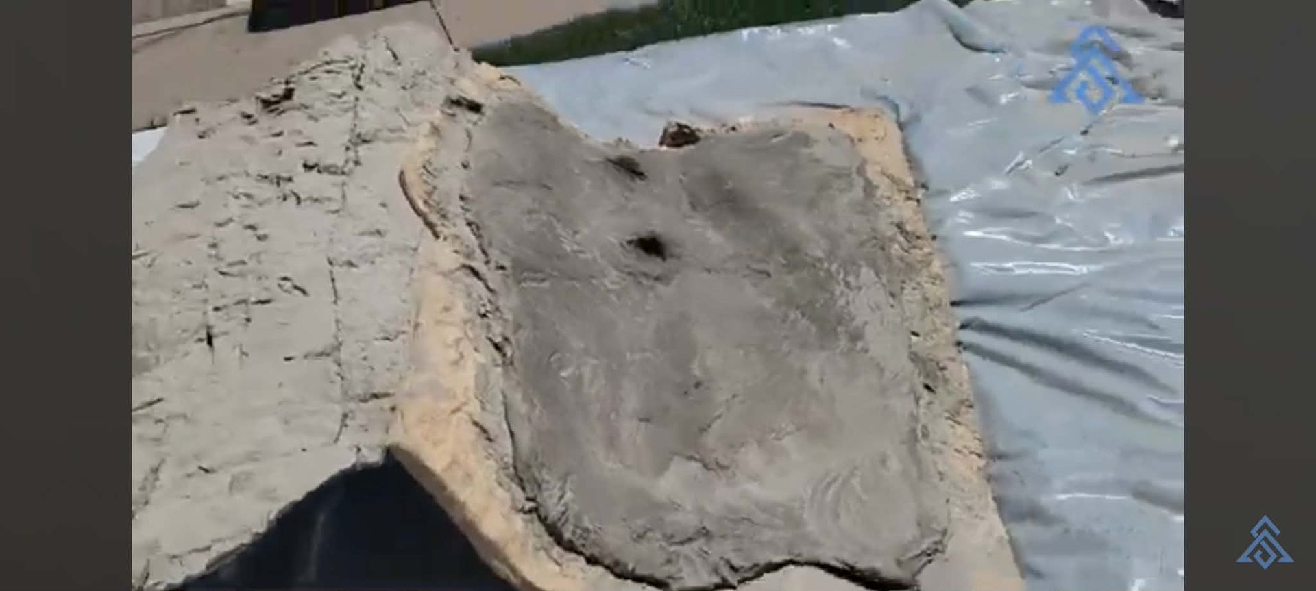 Concrete slab with two holes resting on a blue tarp outdoors, beside a rough stone surface