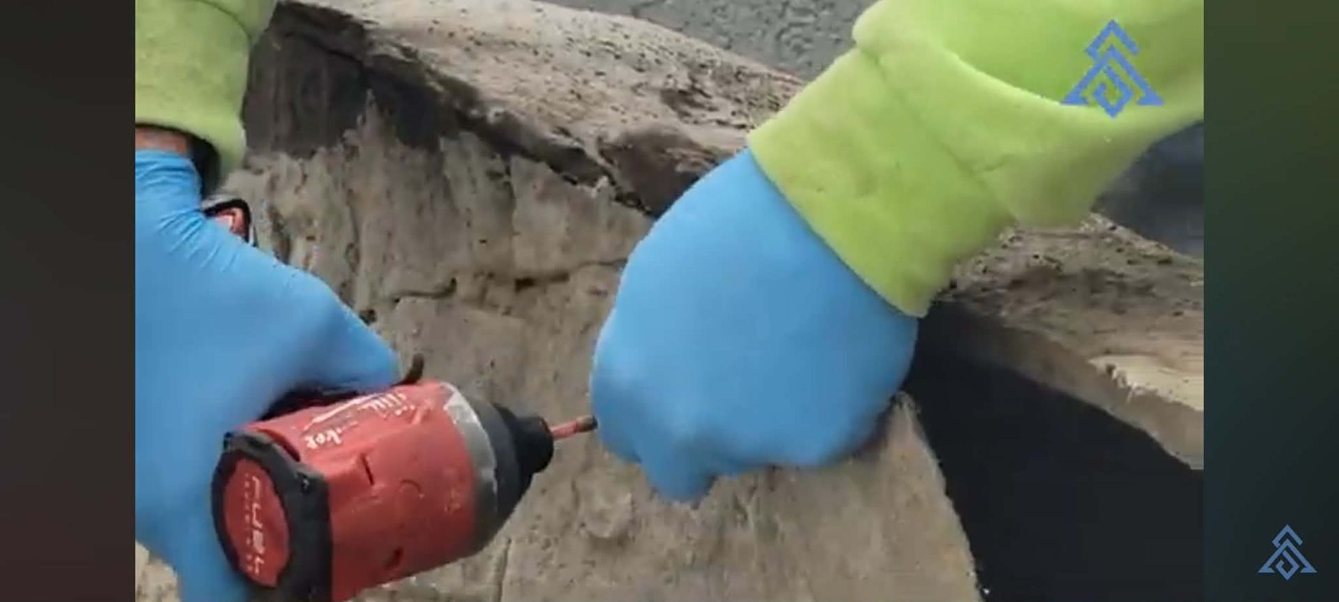 Gloved hands using a red drill to bore into a concrete wall outdoors