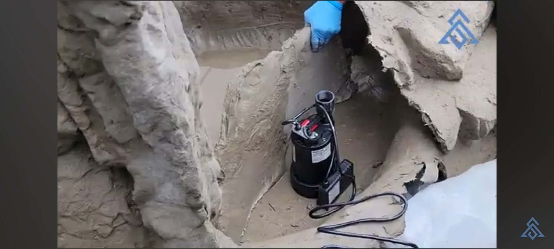 Worker in a narrow trench using a black hose near a concrete wall and wearing a blue glove.