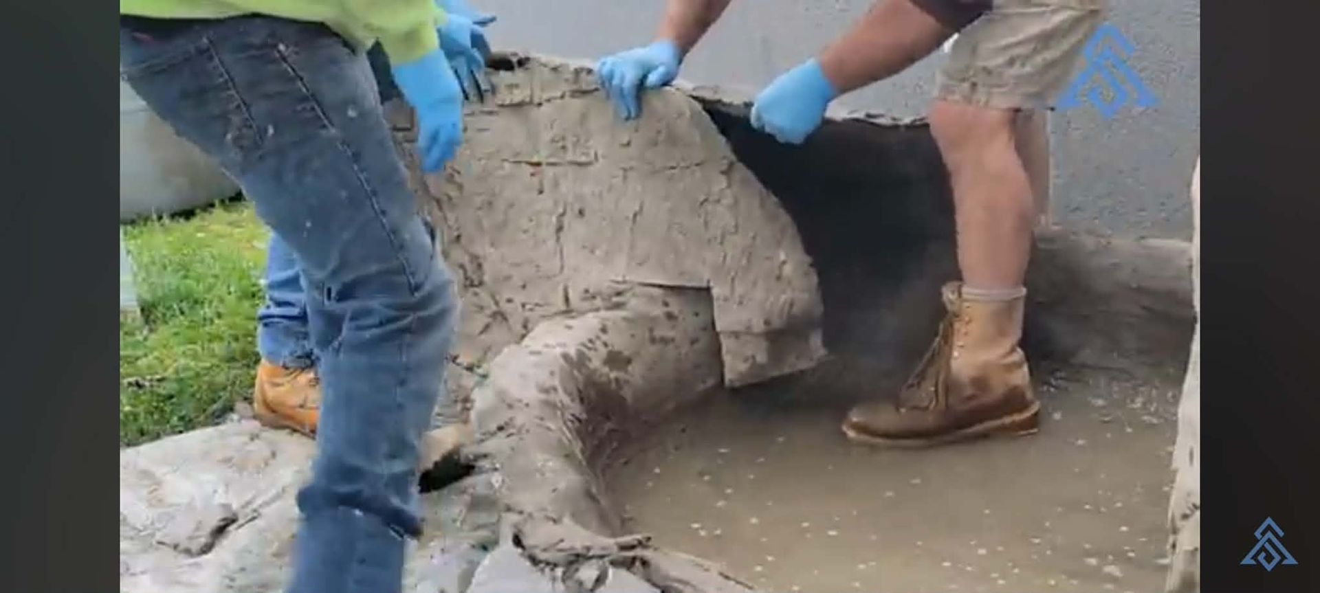 Two workers in gloves lift a large concrete block beside a curb on a wet street.