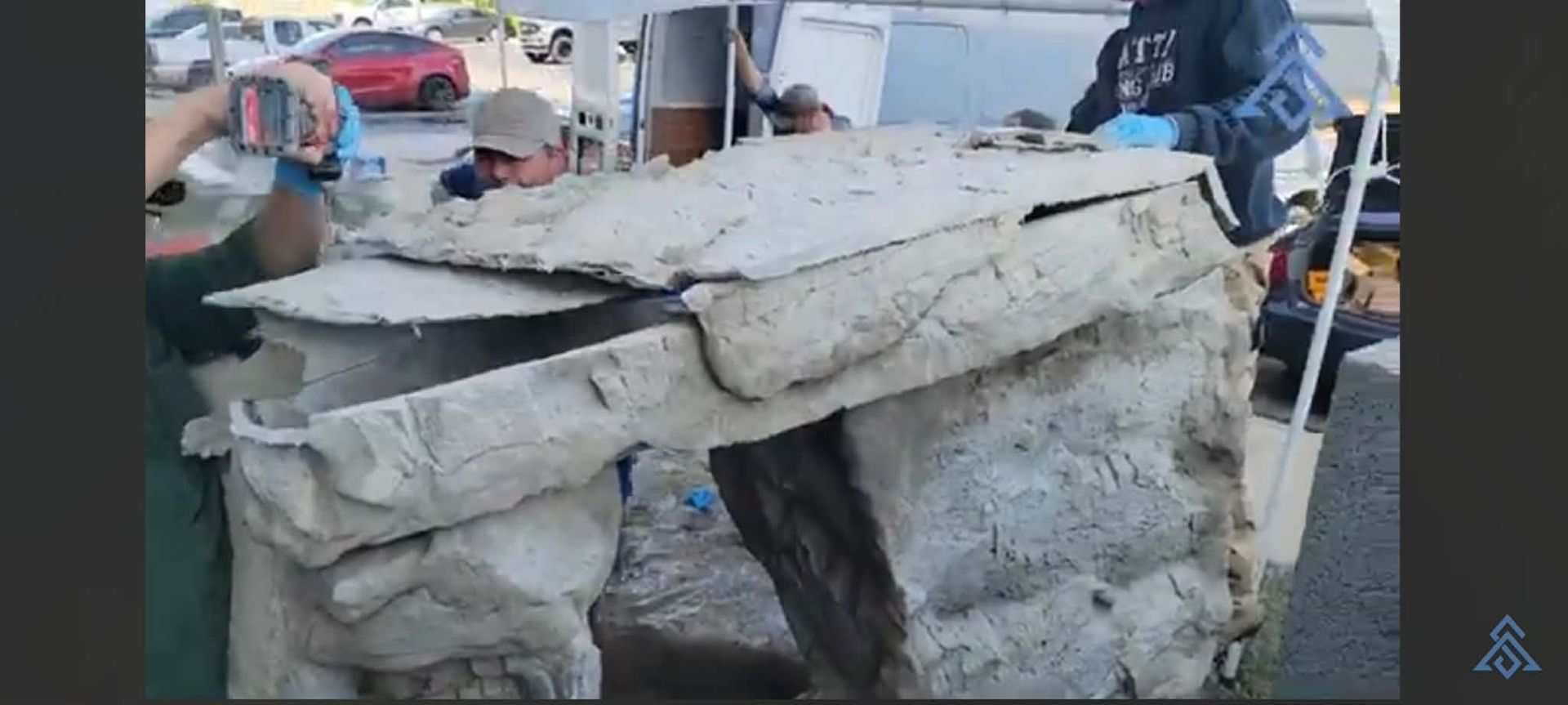 Damaged concrete structure in a yard, with people and parked vehicles in the background.