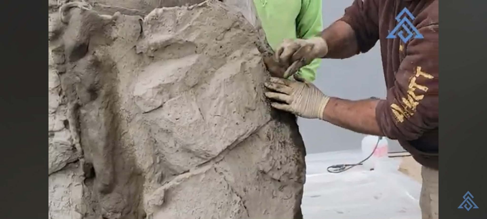 Person shaping wet clay on a large sculpture outdoors, with hands visible in close-up.