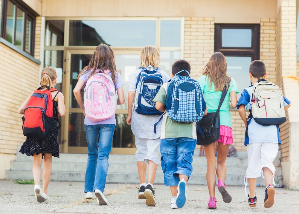 Barn med ryggsekker på vei mot inngangen til en skole i solskinnet