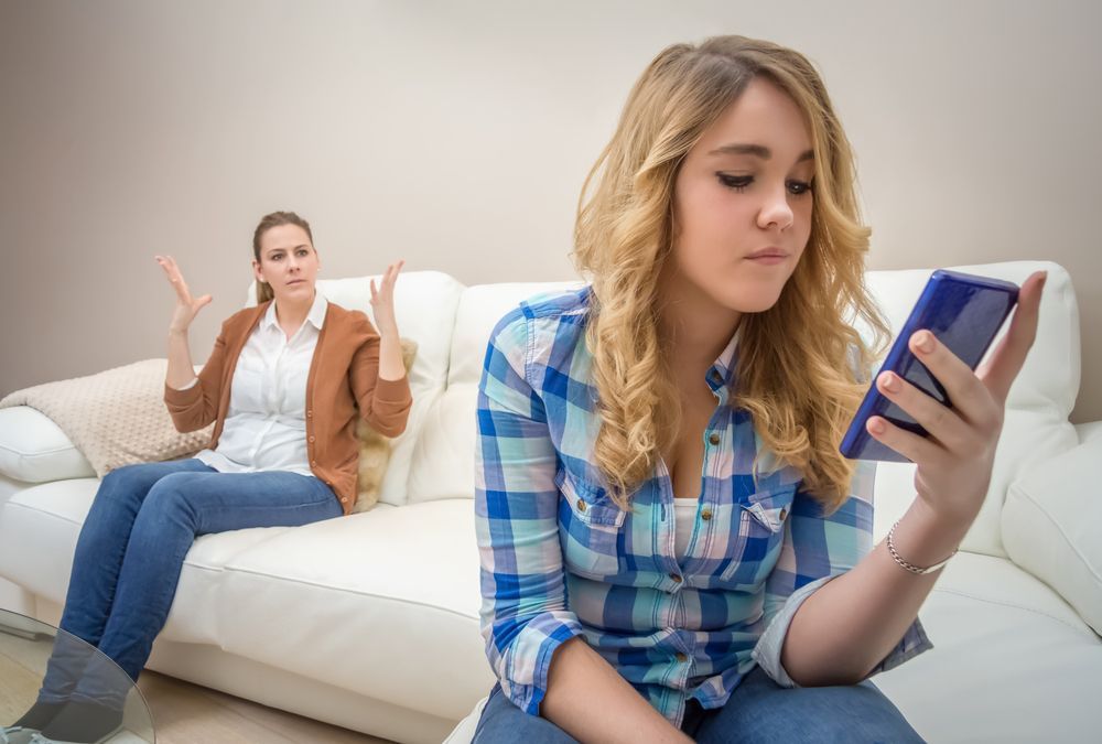Kvinne i blå rutete skjorte ser på telefonen i sofaen; en annen person sitter bak henne og gestikulerer i en stue.