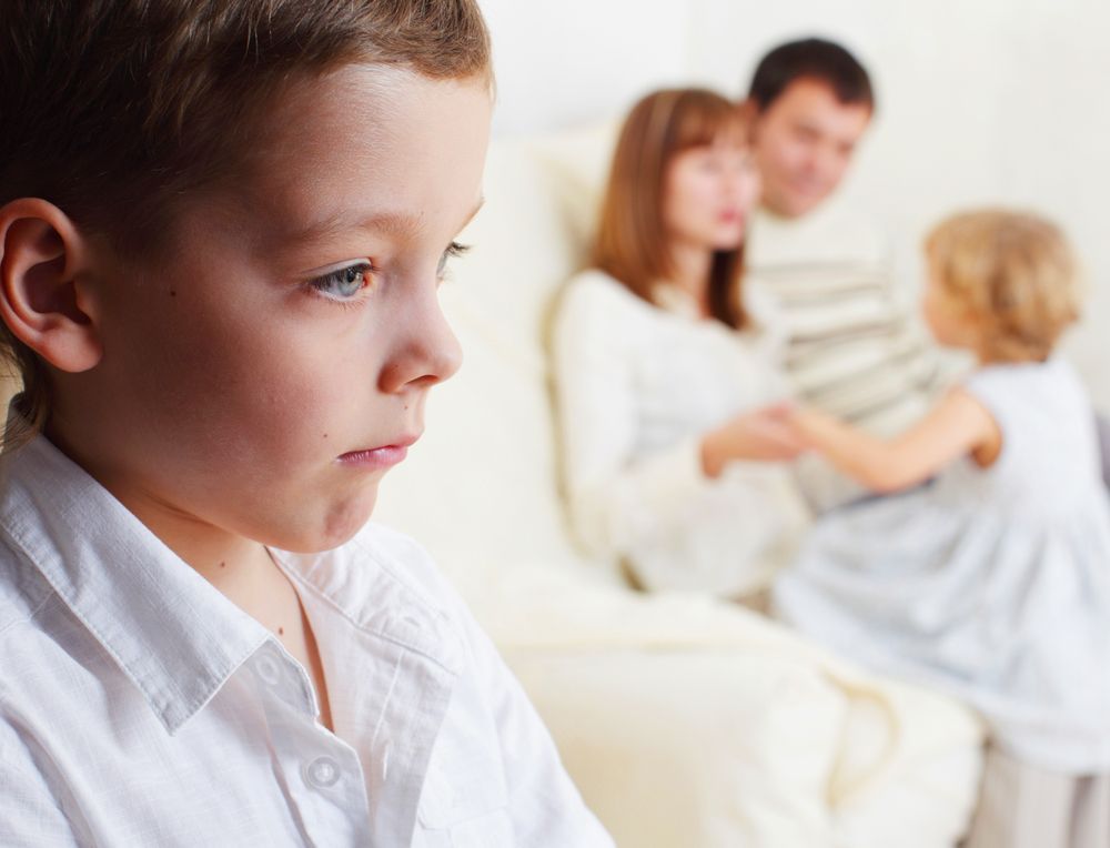 Et lite barn i en hvit skjorte ser til siden, mens en uskarp familie sitter sammen i bakgrunnen.