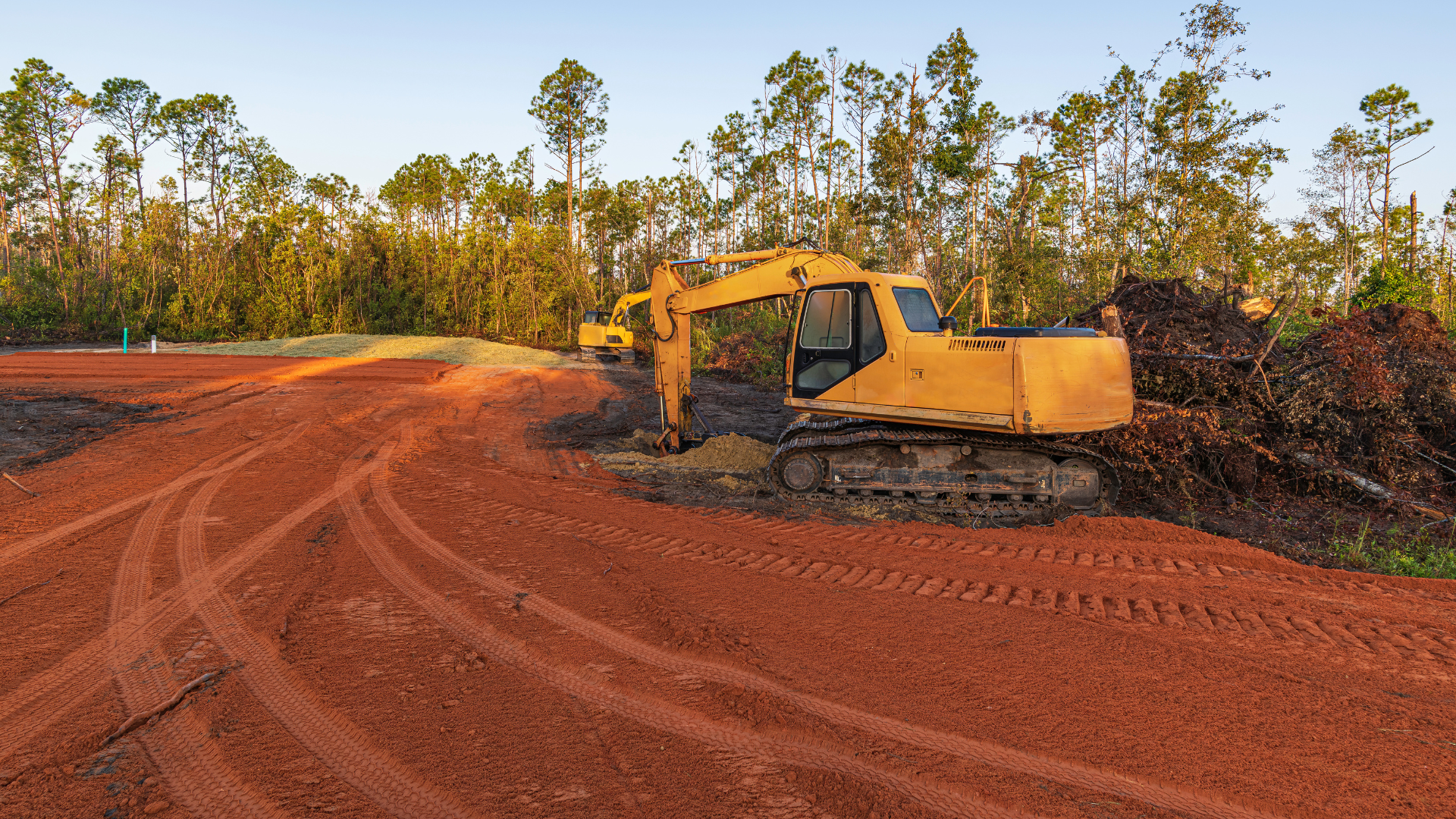 A yellow excavator on a red dirt site next to a pile of brush, with a forest line in the background.