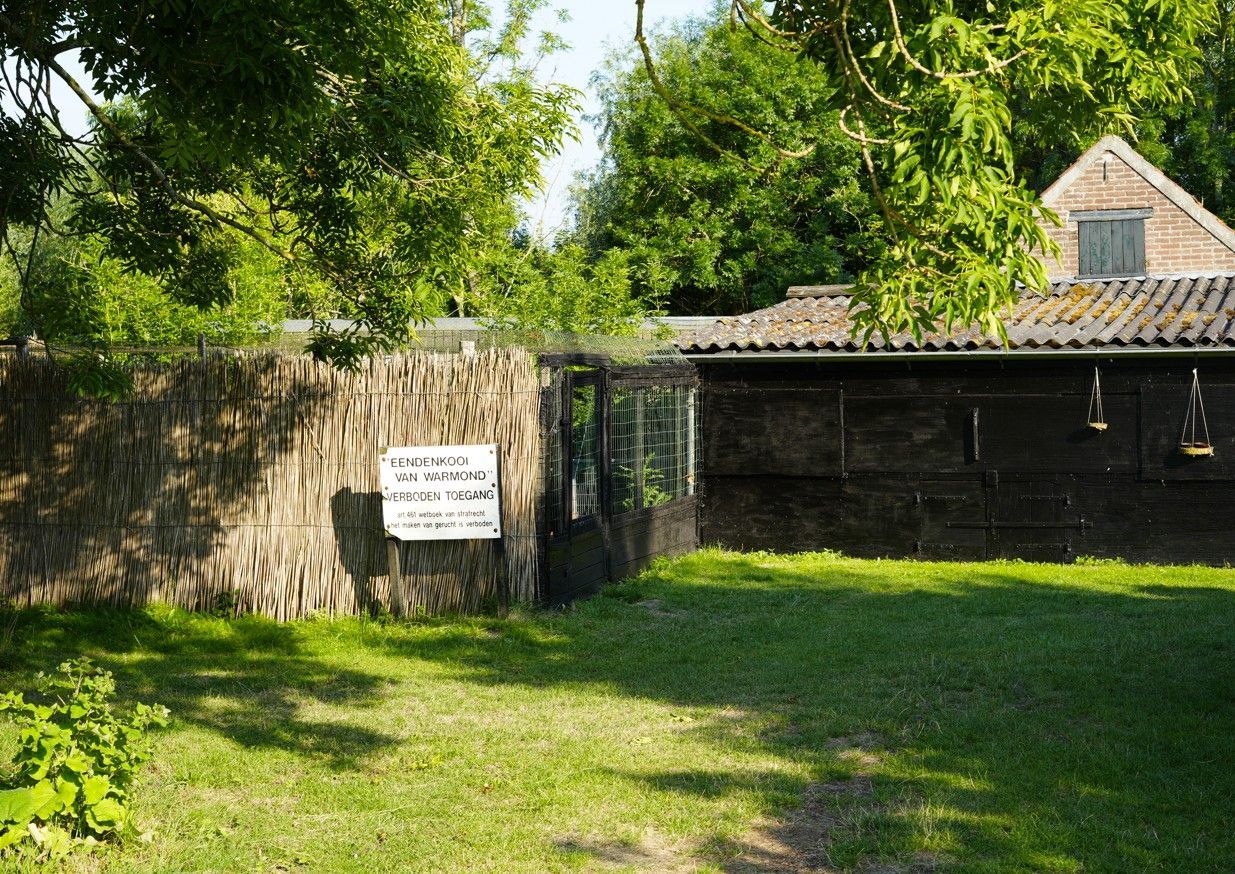 Een verweerd gebouw met een groen gazon. Aan het gebouw is een bord bevestigd. Op de achtergrond zijn bomen te zien.