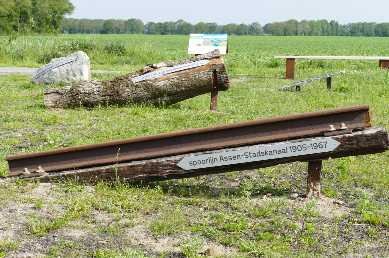 Roestende spoorrails en boomstammen op een grasveld, met in de verte een bord en bomen.
