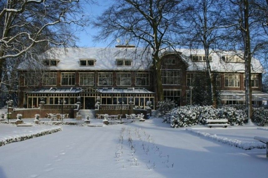 Met sneeuw bedekt gebouw van twee verdiepingen met grote ramen en een overdekte veranda; omringd door bomen.