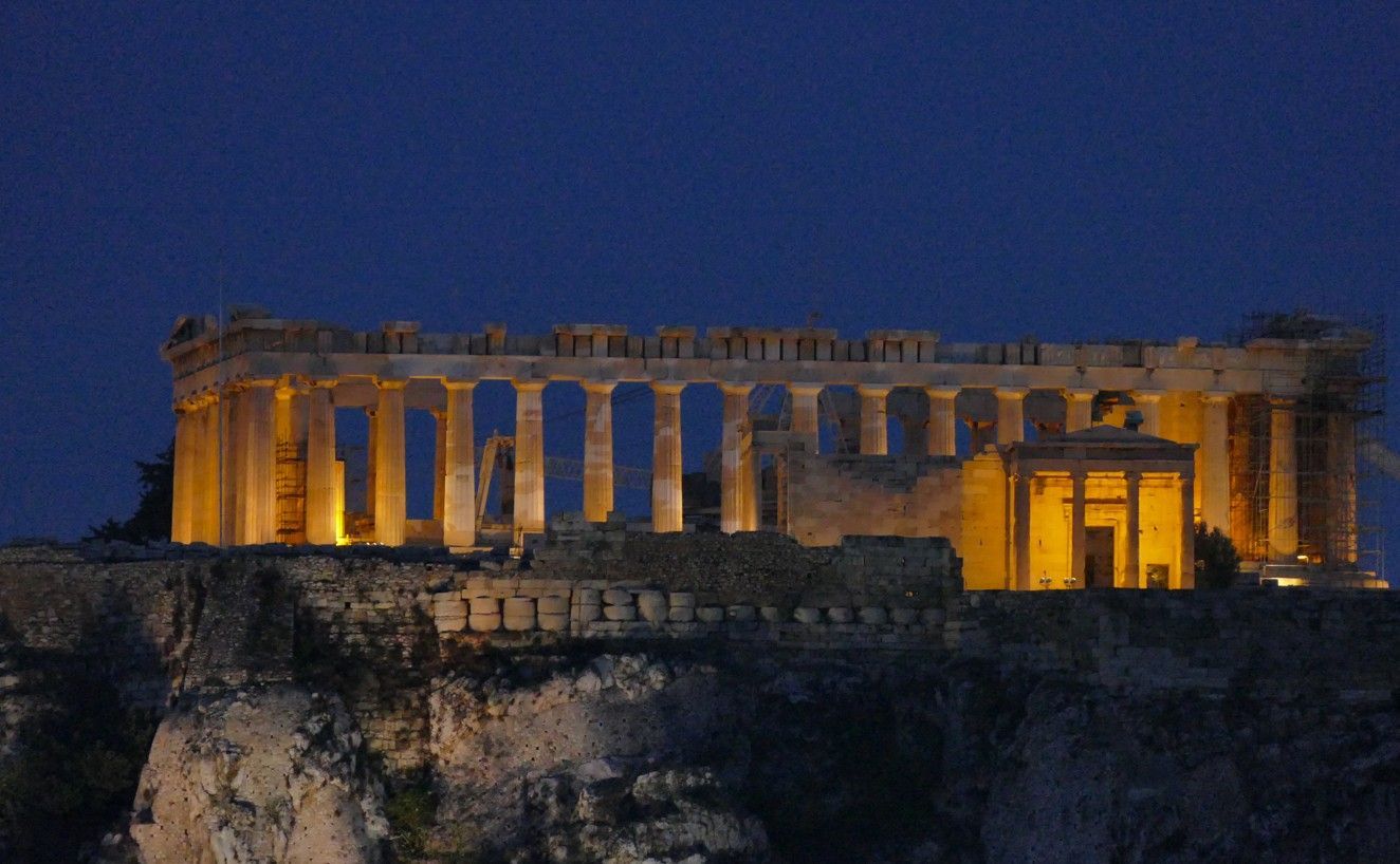 Het Parthenon, 's nachts verlicht, staat bovenop een rotsachtige heuvel tegen een donkerblauwe hemel.