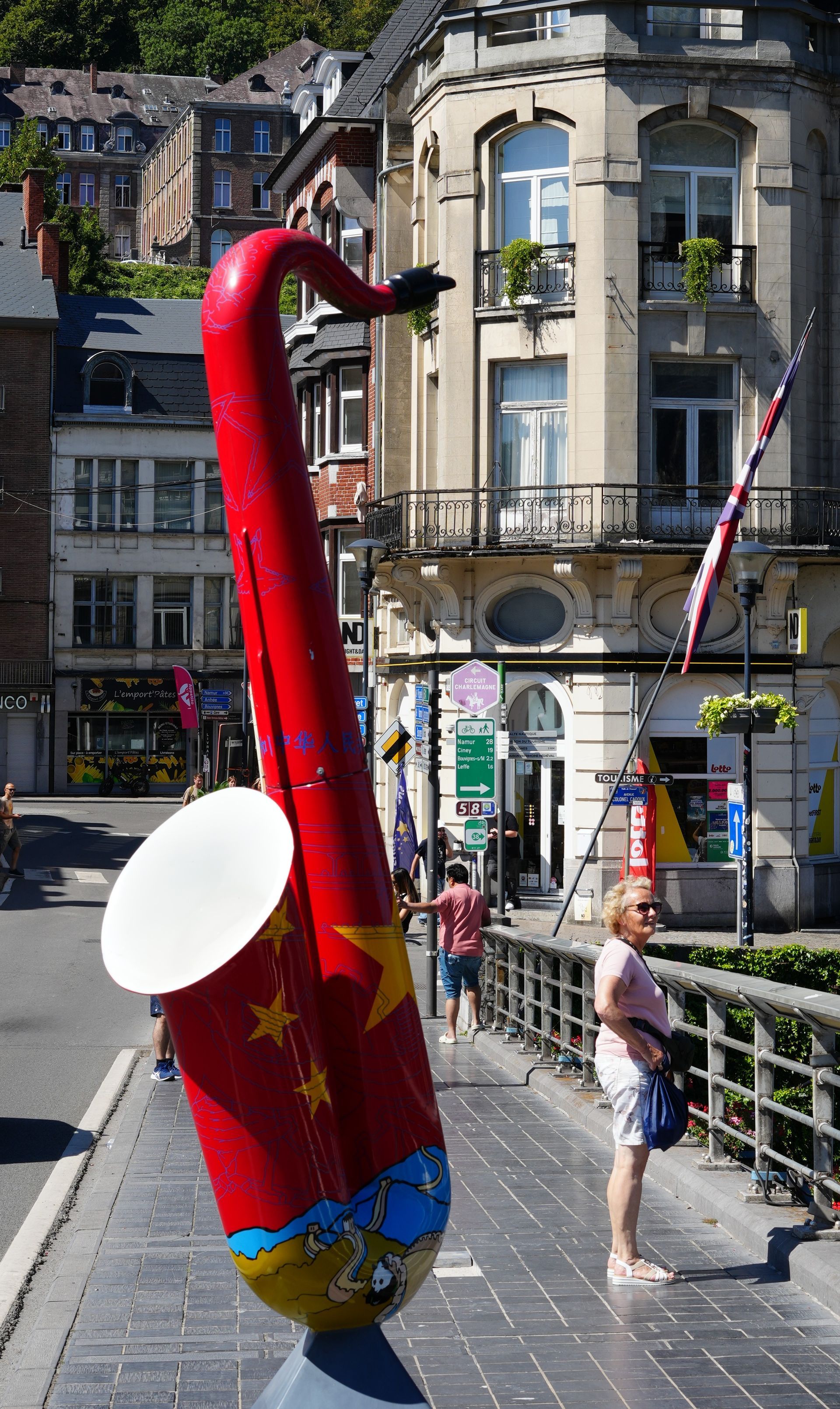 Een gigantisch rood-blauw saxofoonbeeld op een stoep in de stad, vlakbij een vrouw en historische gebouwen.