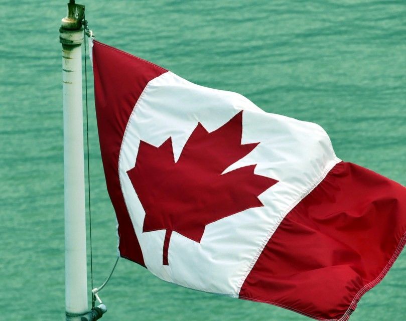 De Canadese vlag wappert aan een vlaggenmast, met een rood esdoornblad op een witte achtergrond met rode randen, tegen een blauwgroene achtergrond.