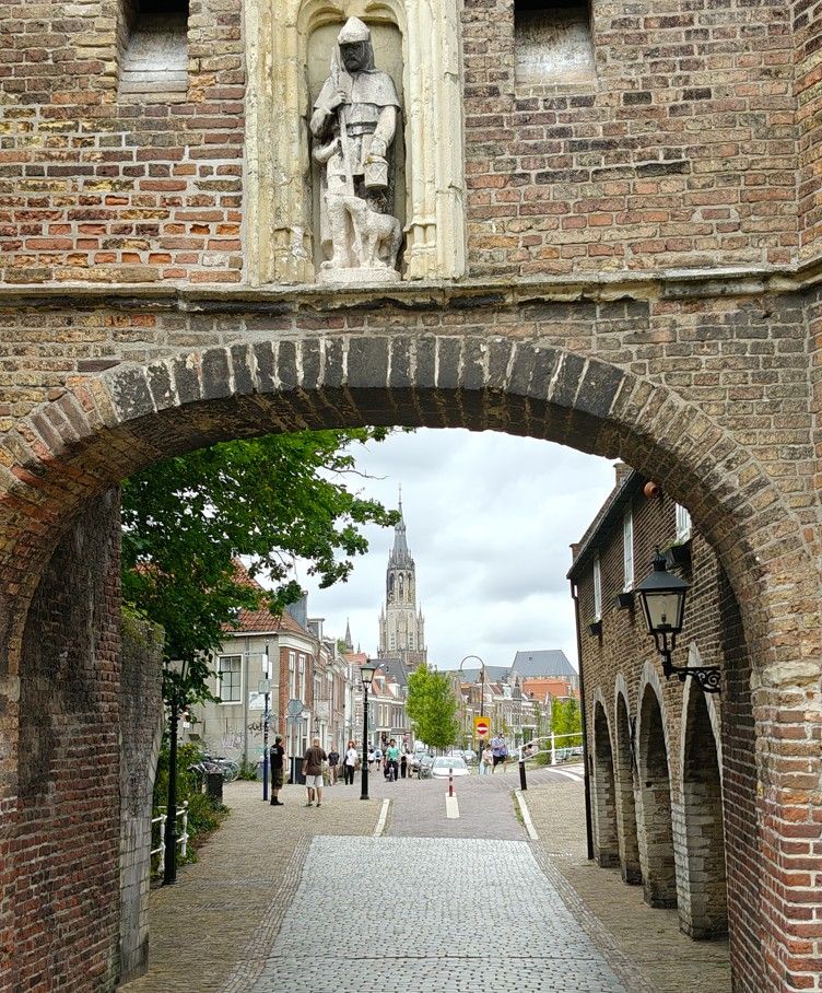Een bakstenen boog omlijst een straat met kinderkopjes die leidt naar een kerktoren in de verte in een historische stad.