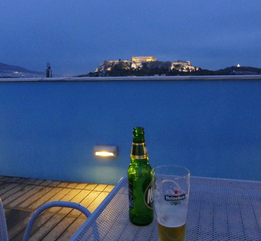 Een bierfles en een glas op een terras met uitzicht op de verlichte Akropolis in Athene bij schemering.