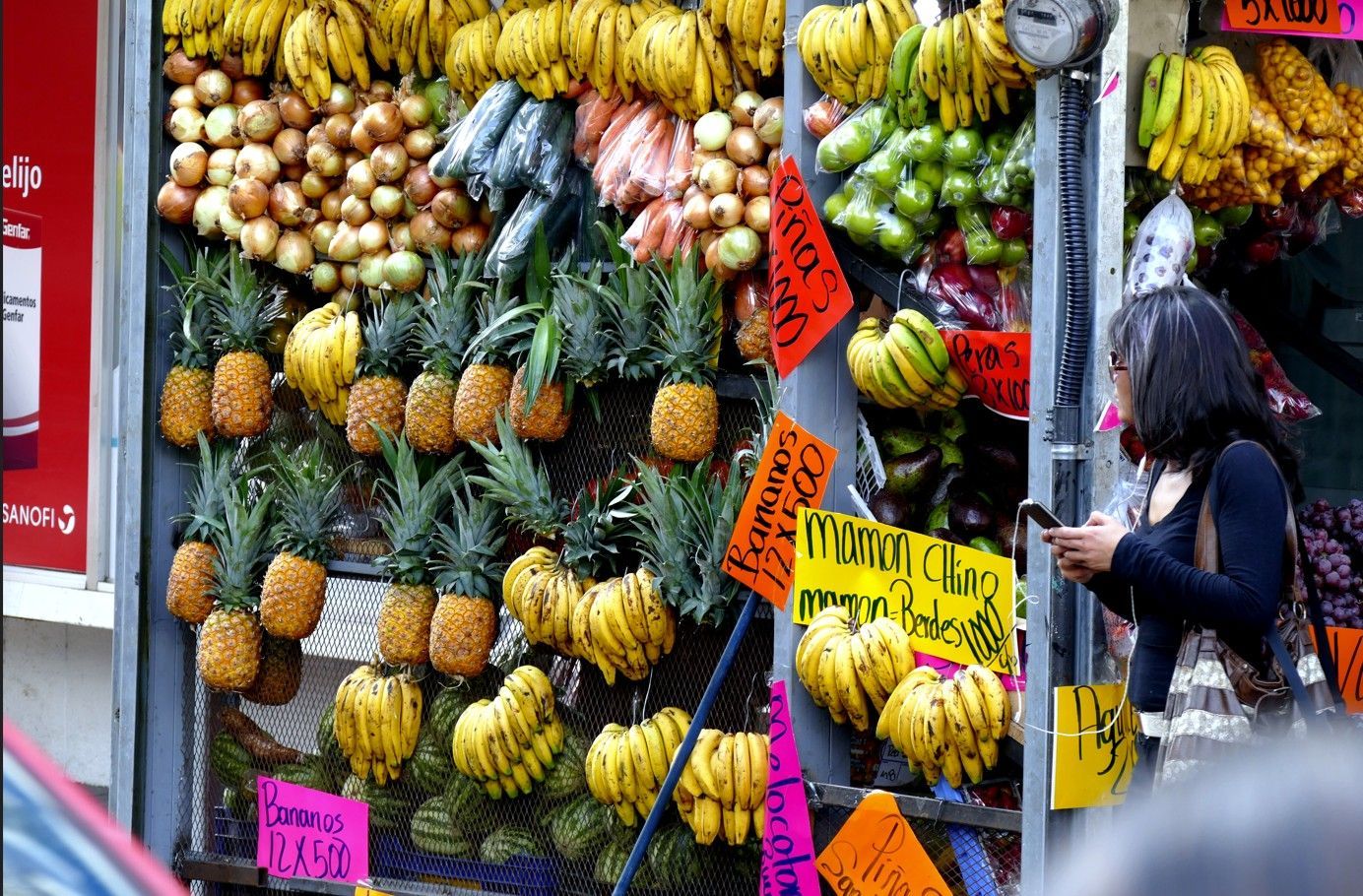 Een fruitkraam met diverse soorten fruit, een vrouw die in de buurt op haar telefoon kijkt.