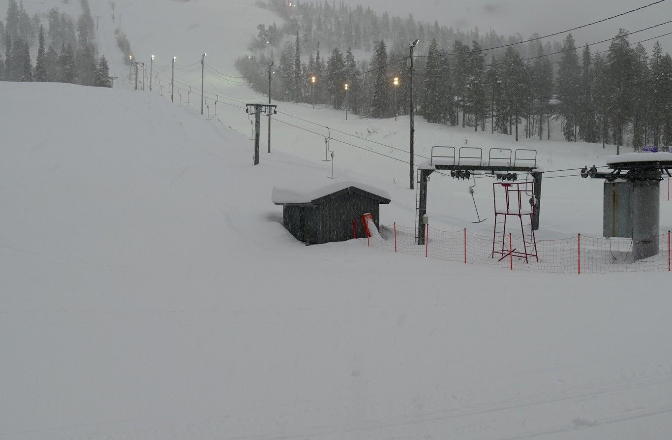 Besneeuwde skipiste met stoellift, klein gebouw en bomen; bewolkte dag.