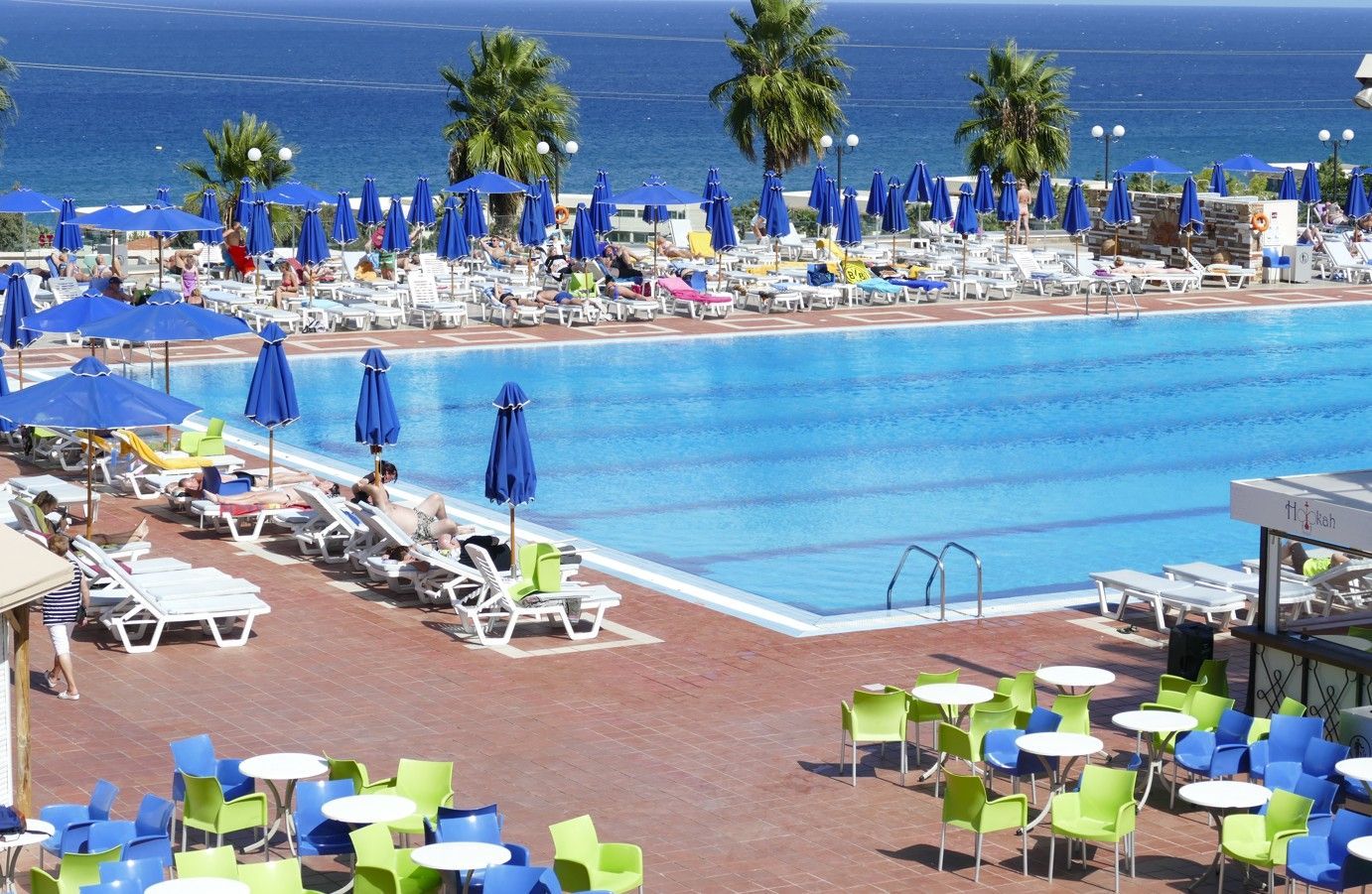 Een tafereel bij het zwembad met een blauw zwembad, de oceaan, parasols, zonaanbidders en terrastafels.