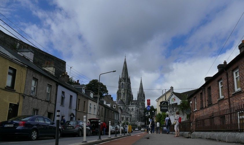 Straatbeeld in Cobh, Ierland, met de kathedraal in de verte, geflankeerd door kleurrijke gebouwen en een bewolkte hemel.