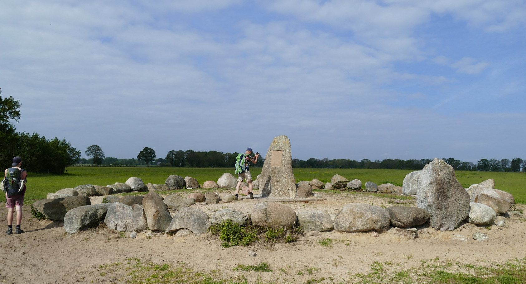 Een stenen cirkelmonument in een grasveld onder een gedeeltelijk bewolkte hemel, met een paar mensen in de buurt.