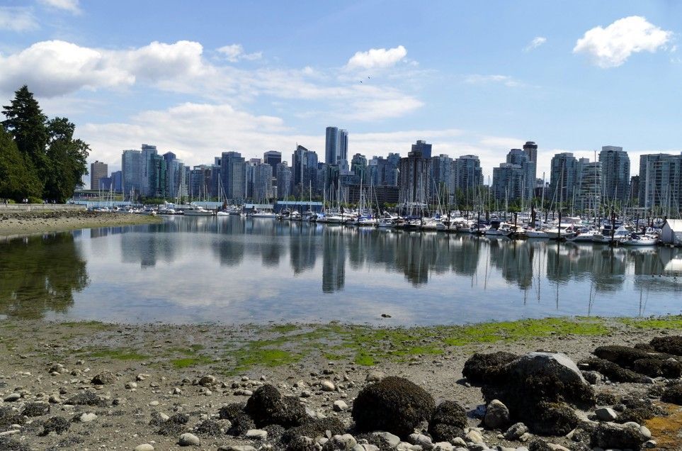 De skyline van Vancouver met op de voorgrond aangemeerde boten, weerspiegeld in het kalme water onder een blauwe hemel.