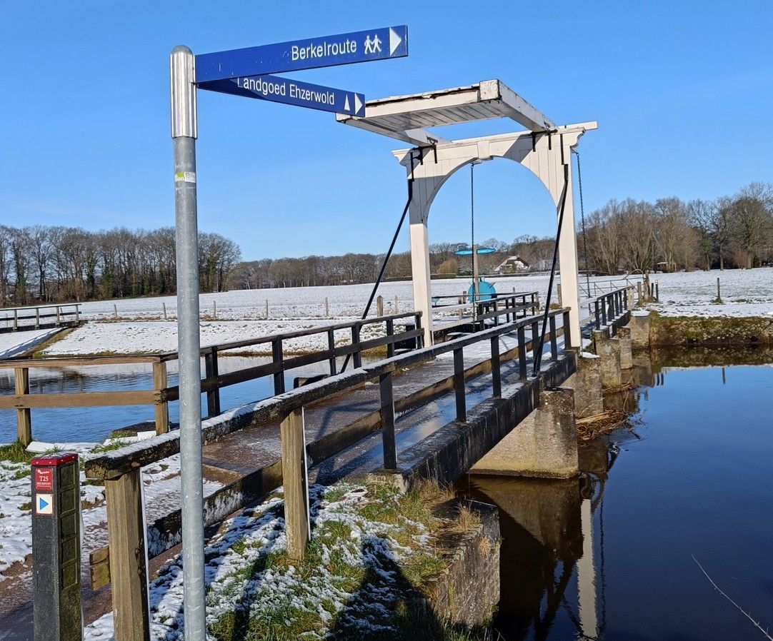 Een brug over een kanaal met besneeuwde oevers en een blauw bord.