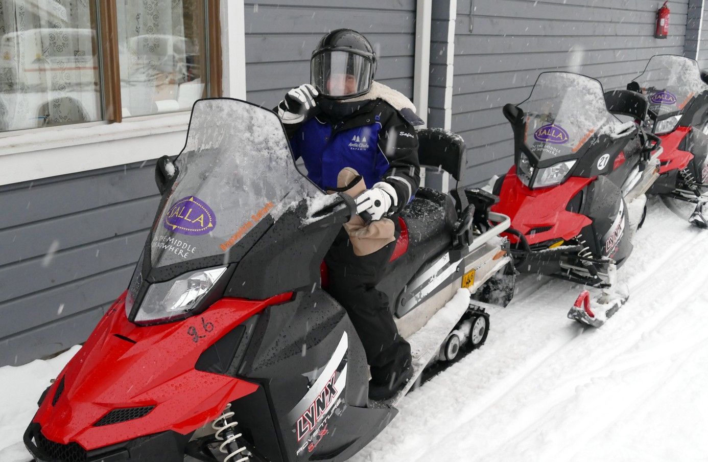 Persoon op een rood-zwarte sneeuwscooter, anderen in de rij. Besneeuwde omgeving.