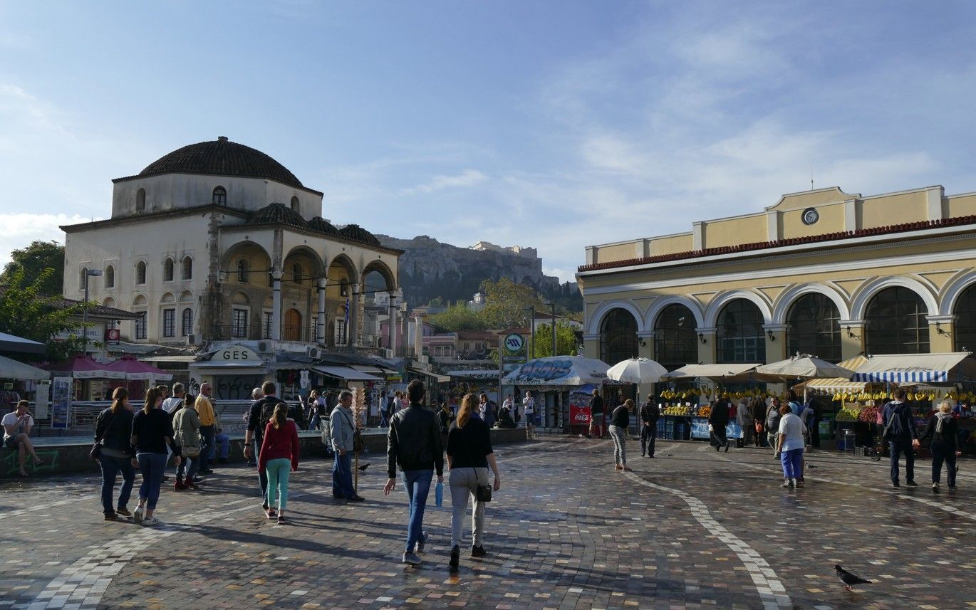Een levendig plein in Athene met een historische moskee en markt; mensen wandelen en winkelen onder een blauwe hemel.