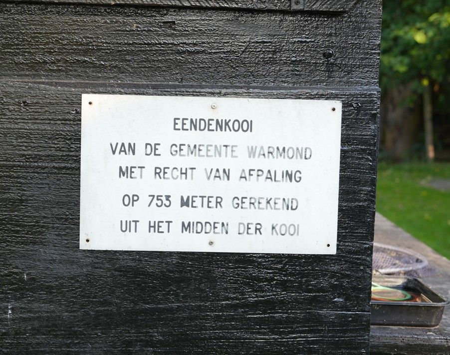 White rectangular sign on a dark wall. Text in Dutch: “EENDENKOOI VAN DE GEMEENTE WARMOND...755 meter...”