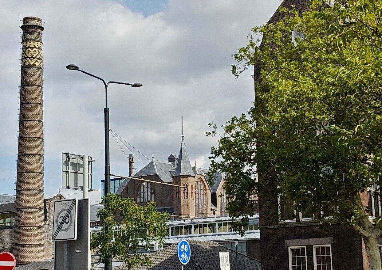 Straatbeeld met een hoge schoorsteen, een historisch gebouw en bomen onder een gedeeltelijk bewolkte hemel.