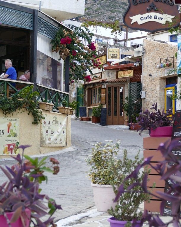 Smalle straat met kinderkopjes, uithangborden van cafés en potplanten. Mensen zitten aan tafels buiten.