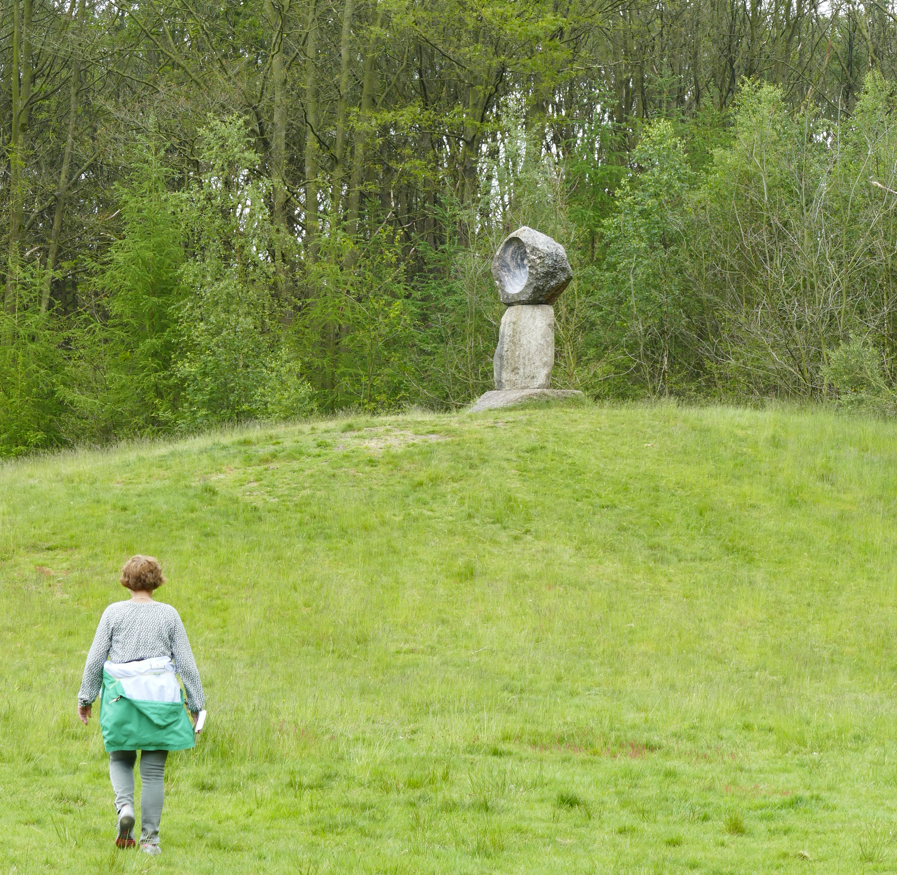 Een persoon loopt een grasheuvel op richting een stenen sculptuur in een groen, bebost park.