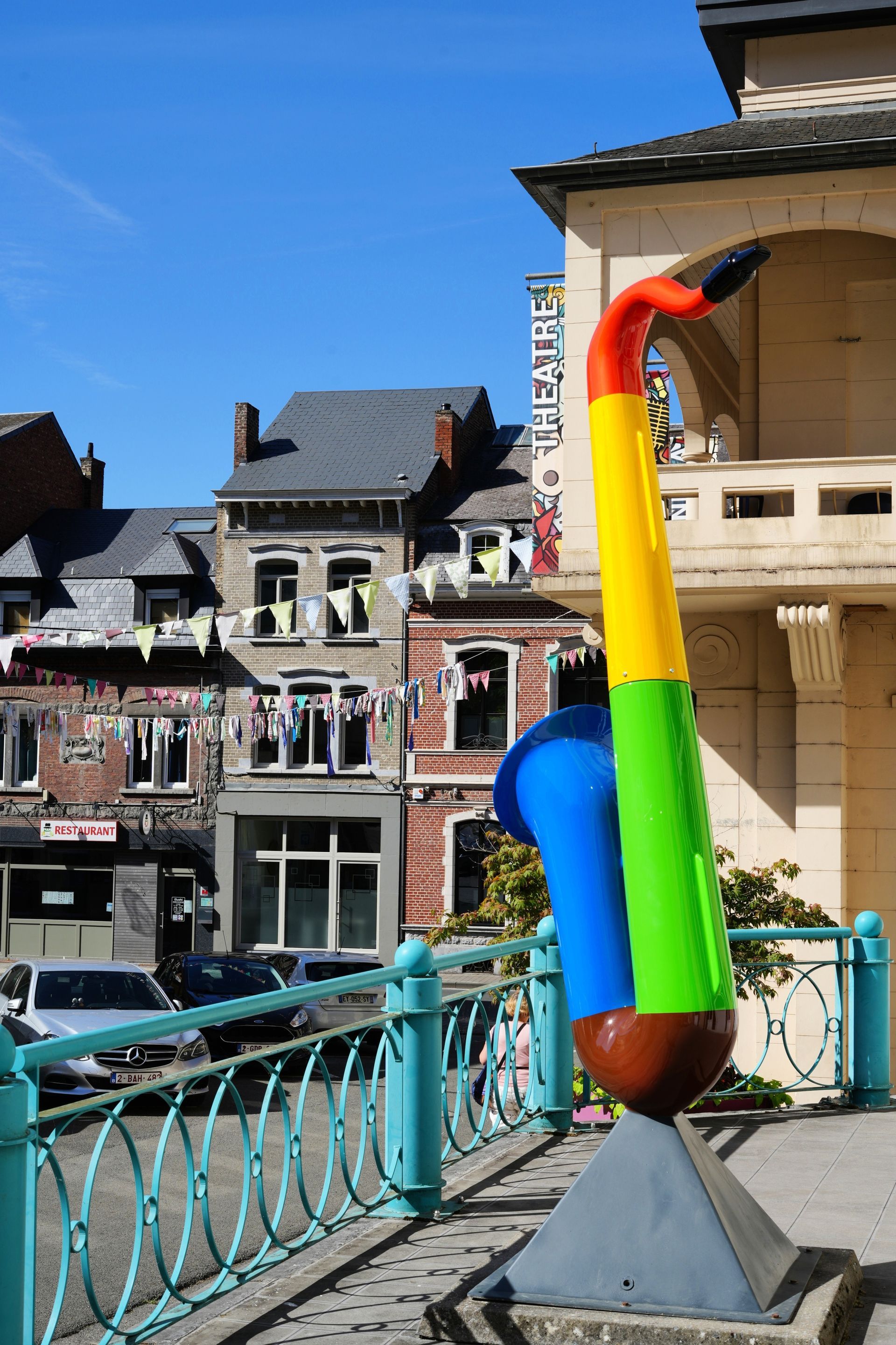 Een kleurrijk abstract beeldhouwwerk op een brug met uitzicht op een historische straat onder een blauwe hemel.