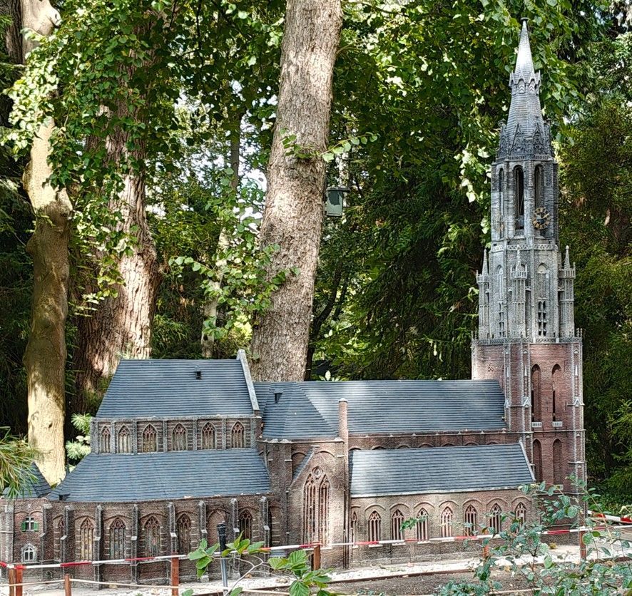 Miniatuur bakstenen kerkje met hoge torenspits te midden van bomen in een bosrijke tuin.