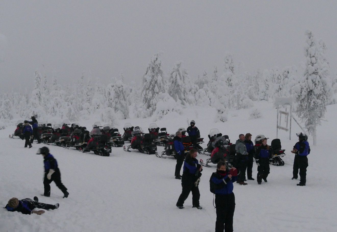 Een groep mensen in blauwe jassen en sneeuwkleding bij sneeuwscooters in een besneeuwd landschap.