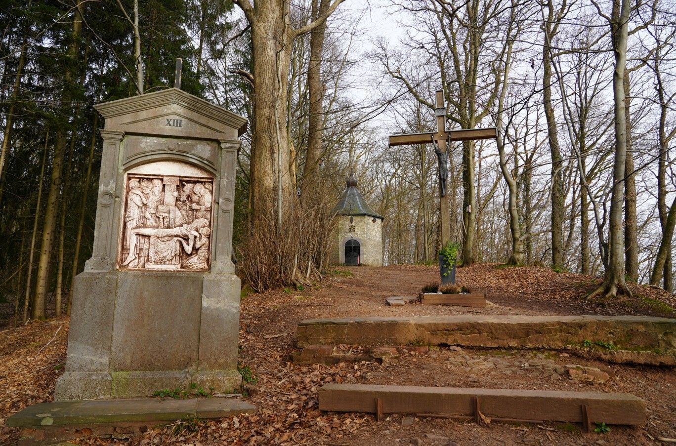 Op een bebost pad dat naar een kleine, ronde kapel leidt, staan ​​een stenen schrijn en een groot houten kruis.