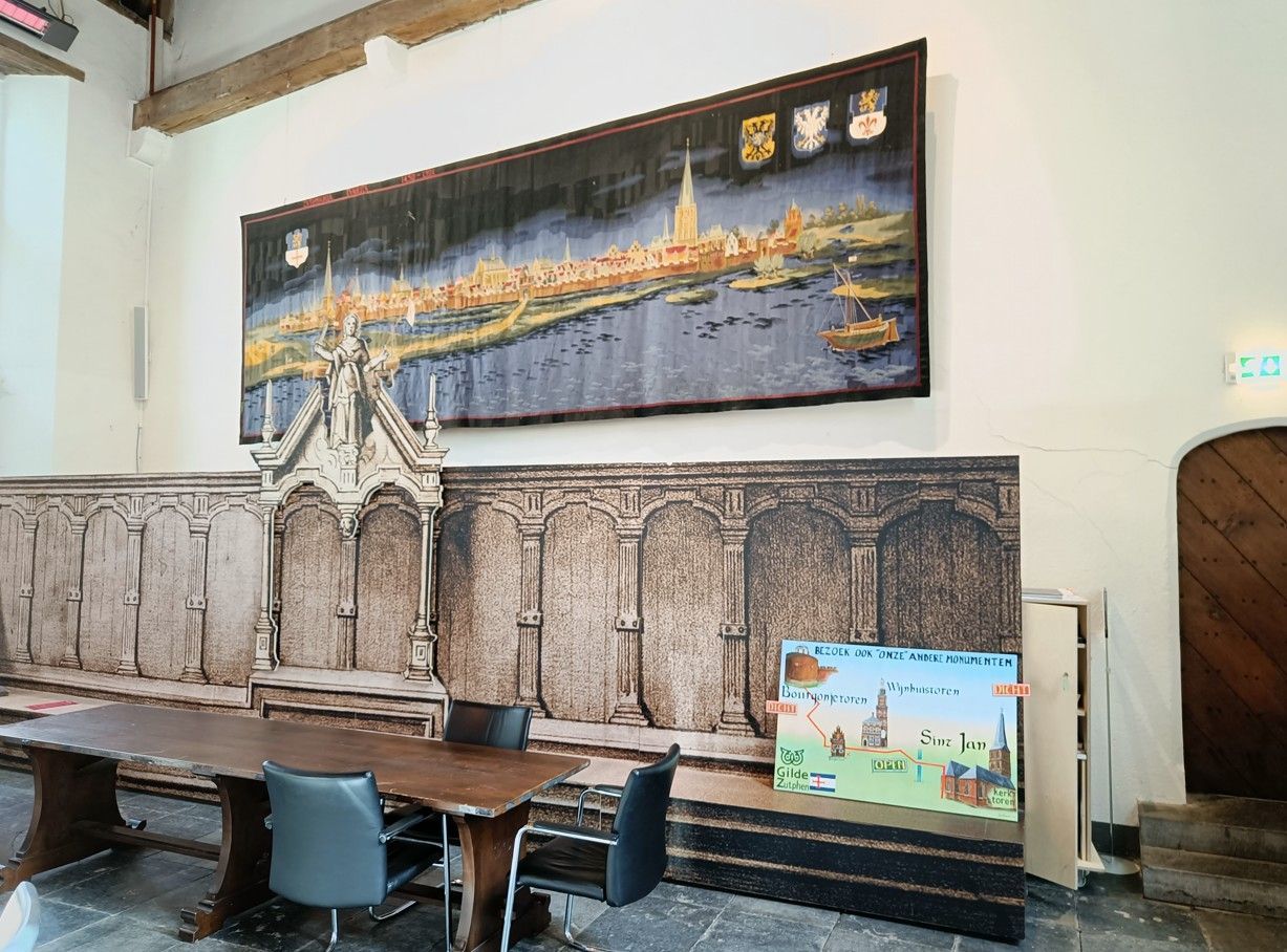 Een groot wandtapijt met een stadsgezicht hangt boven een lange houten tafel en stoelen in een kamer met een decoratieve muur en een houten deur.