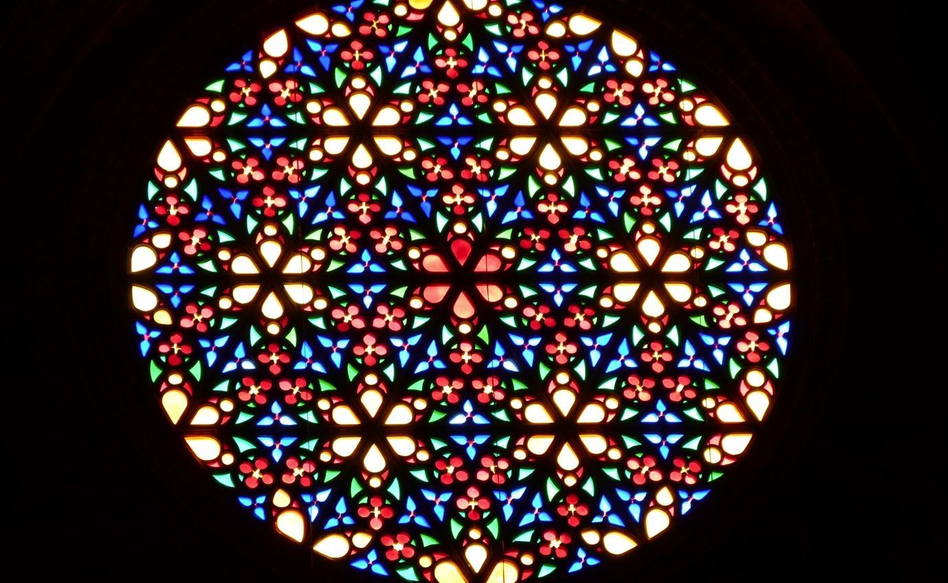 Een roosvenster van gebrandschilderd glas met een geometrisch patroon in rood, blauw, groen en goud.