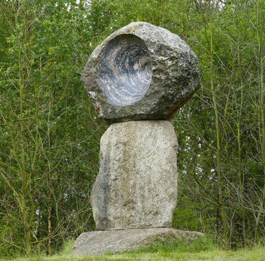 Een stenen sculptuur met een ronde, gebeeldhouwde bovenkant, geplaatst in een grasveld met bomen op de achtergrond.
