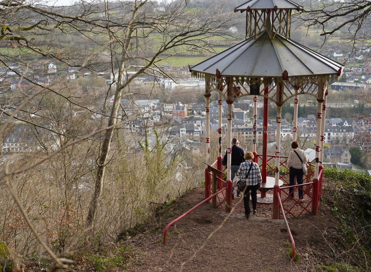 Drie mensen staan ​​in een wit-rood prieel met uitzicht op een stadje in een bosrijk, heuvelachtig landschap.