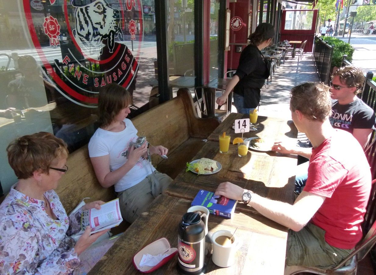 Mensen zitten aan een tafeltje op het terras; eten, drinken, zonlicht.
