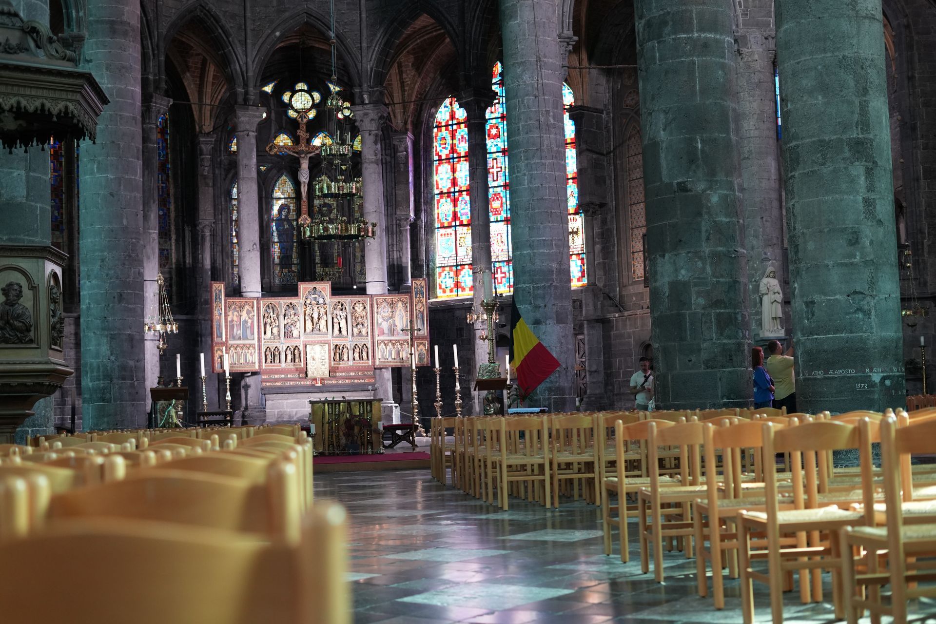 Rijen houten stoelen staan ​​opgesteld tegenover een sierlijk altaar in een schemerige stenen kerk met glas-in-loodramen.