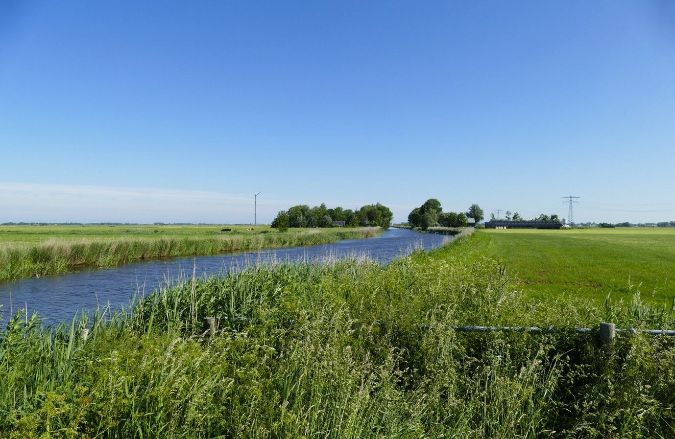 Een kanaal dat door een groene weide stroomt onder een blauwe hemel, met bomen en een gebouw in de verte.