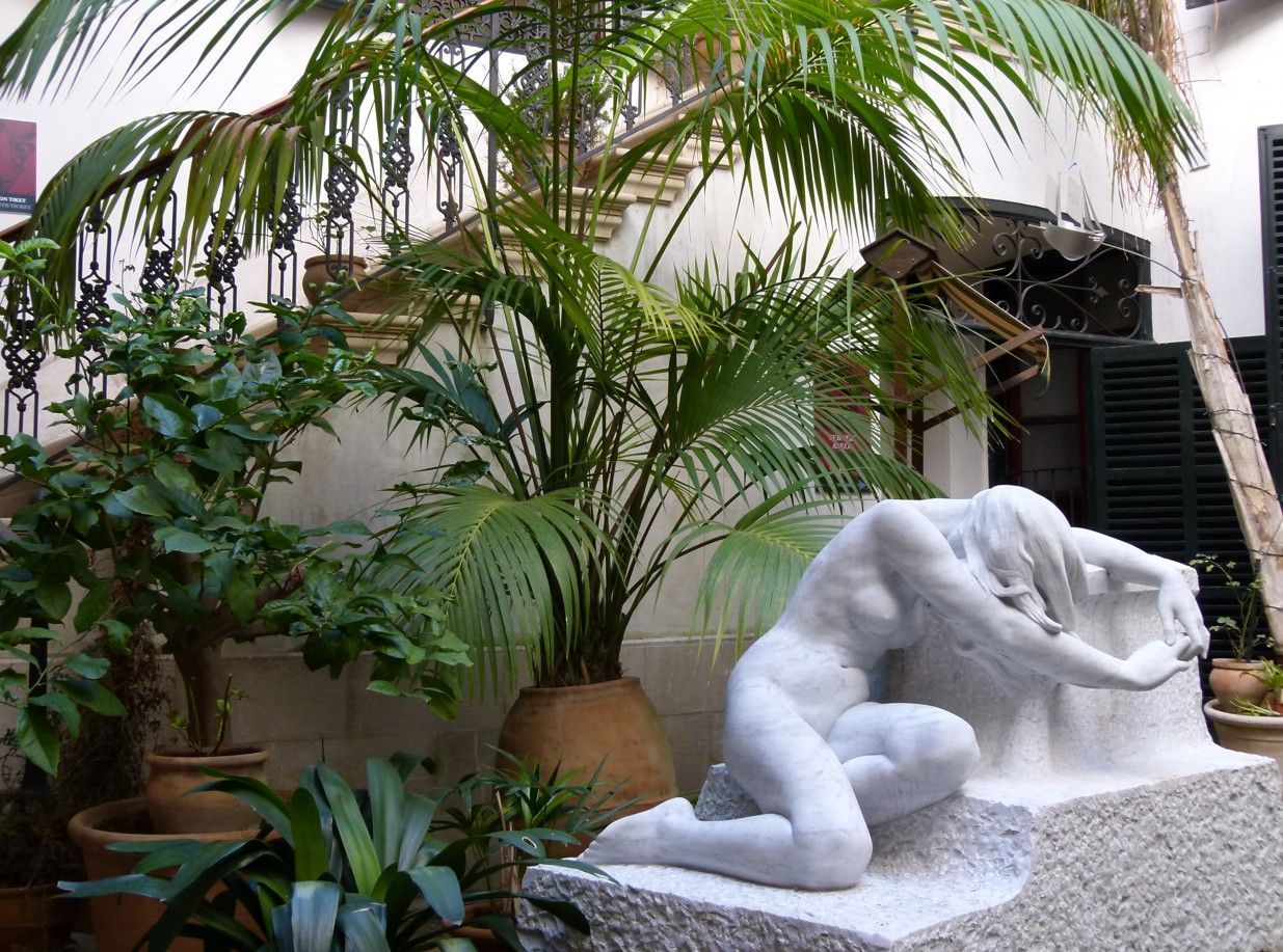 Een wit beeld van een knielende figuur in een binnenplaats met planten.