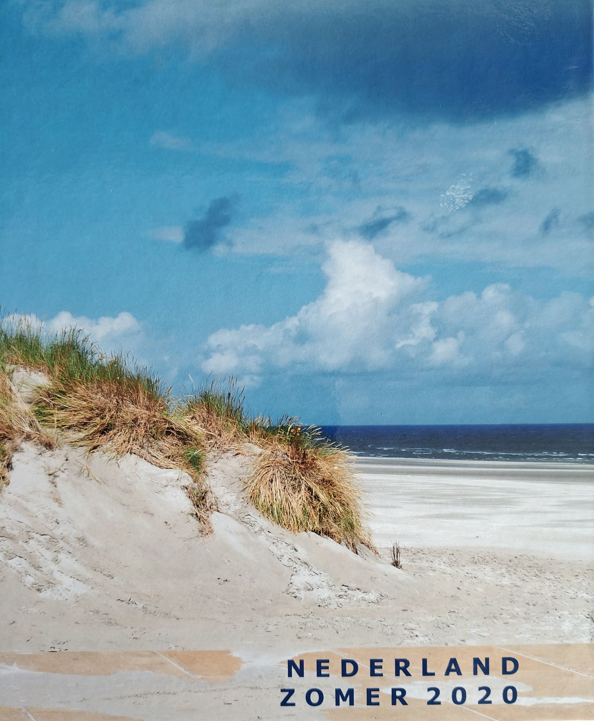 Zandstrand met duinen en oceaan onder een blauwe hemel. Tekst: Nederland Zomer 2020.
