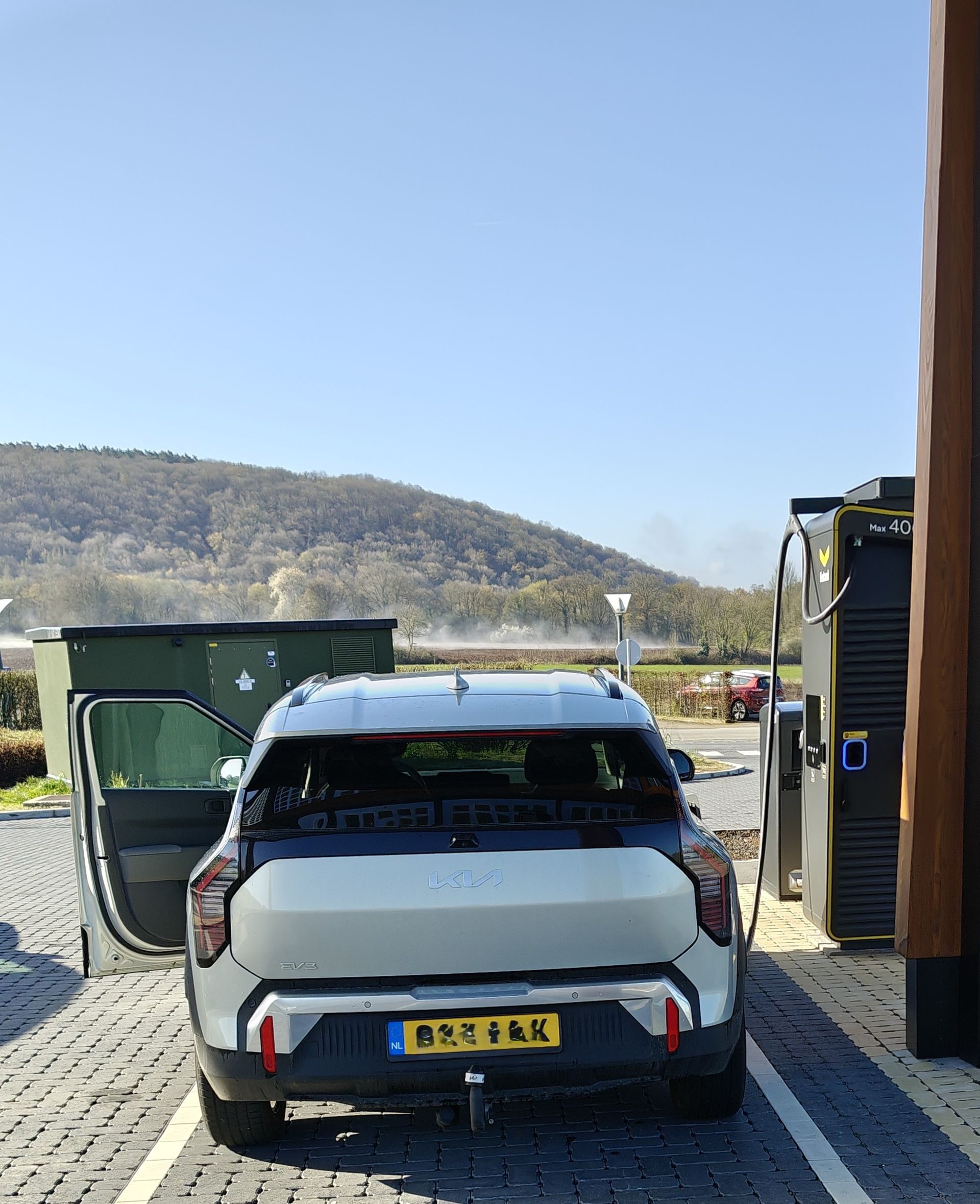 Een zilverkleurige elektrische auto staat geparkeerd bij een laadstation op een zonnige dag.