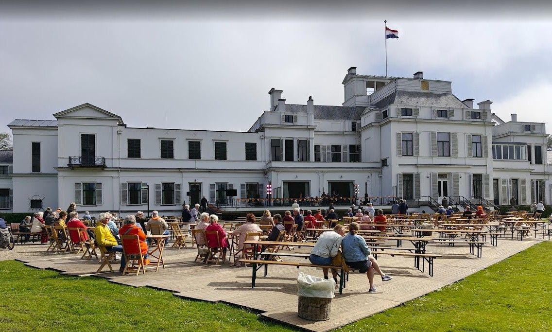 Wit gebouw met mensen die aan tafels buiten zitten, een Nederlandse vlag wappert.